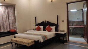 Une chambre à coucher comprenant un lit, un banc, une table de chevet, une fenêtre avec des rideaux et une salle de bain visible à travers une porte ouverte.