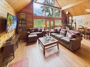 Ein gemütliches Blockhaus-Interieur mit Holzwänden und -böden, einem großen Fenster mit Blick ins Freie, einem Kamin und einem Fernseher.