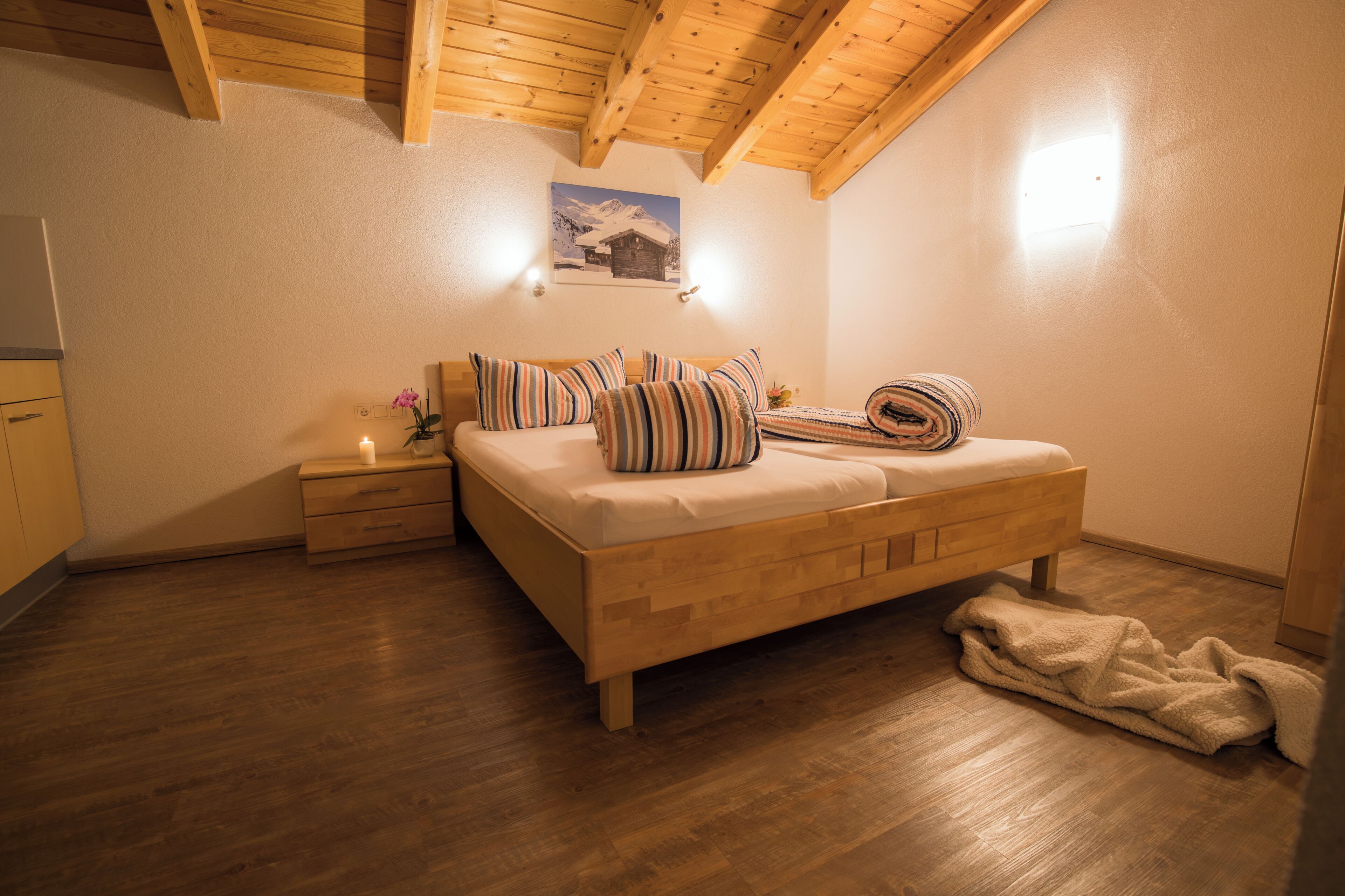 Een slaapkamer met een houten plafond, een bed met gestreept beddengoed, een nachtkastje met een lamp en een ingelijst schilderij aan de muur.