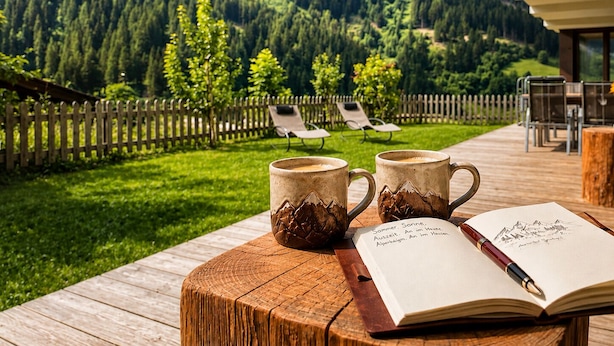 Een houten terras met twee mokken en een boek, met uitzicht op een berglandschap met een houten hek en lounge stoelen.