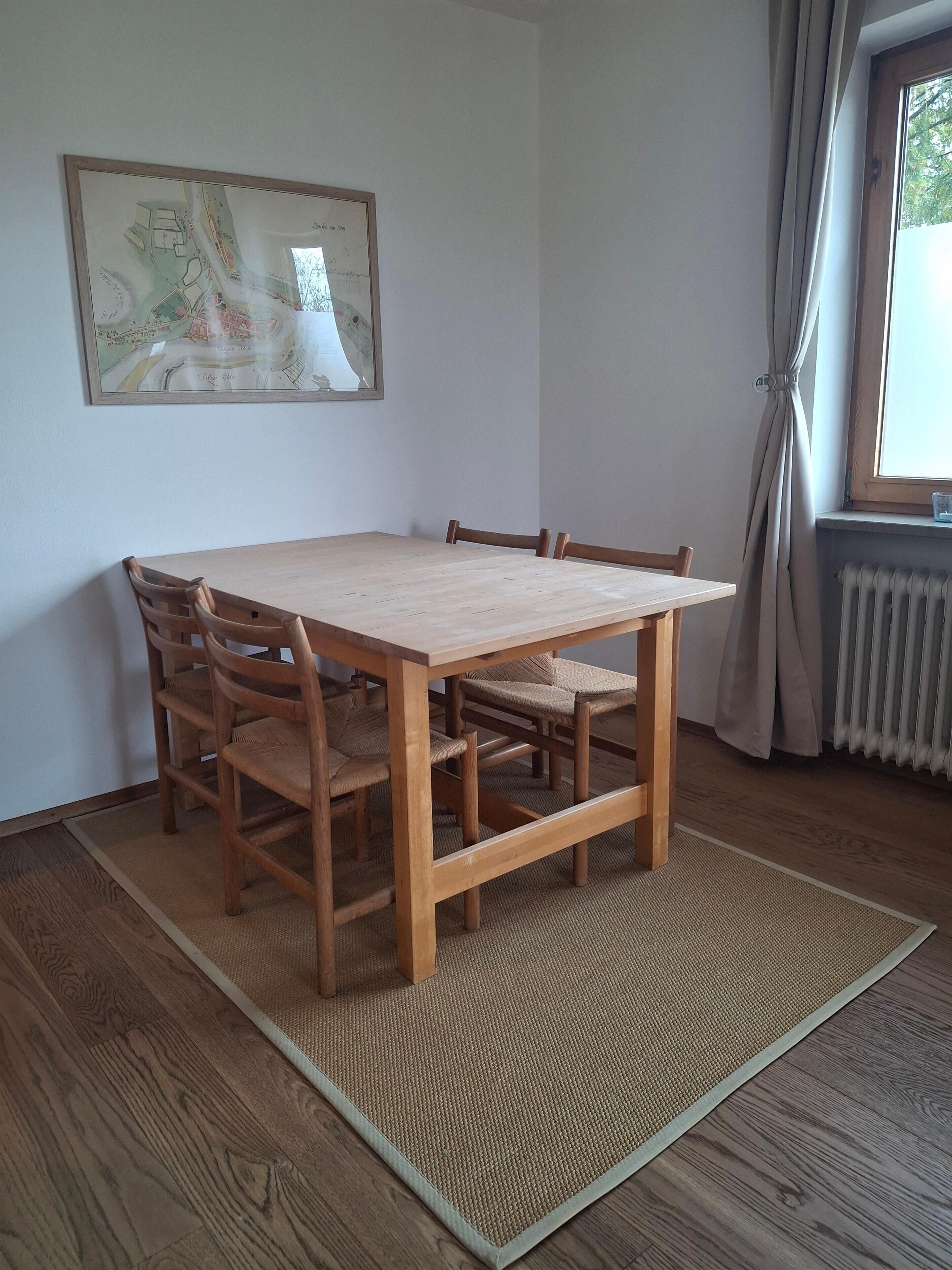 A dining table with chairs, a woven rug, a framed map on the wall, and a window with curtains.