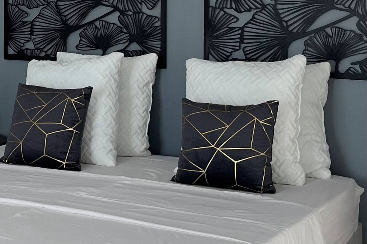 A bed with white bedding, two black wall art pieces with leaf patterns, and two black pillows with gold geometric designs.