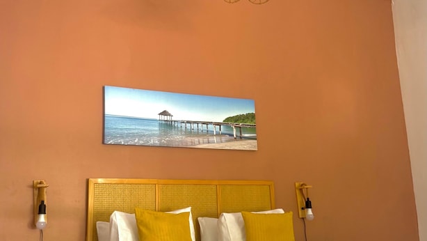 A bed with a white and yellow bedspread, two bedside tables with lamps, a painting of a pier on the wall, and a wooden floor. - La Kaz by Kaz Kafe (Pamandzi)