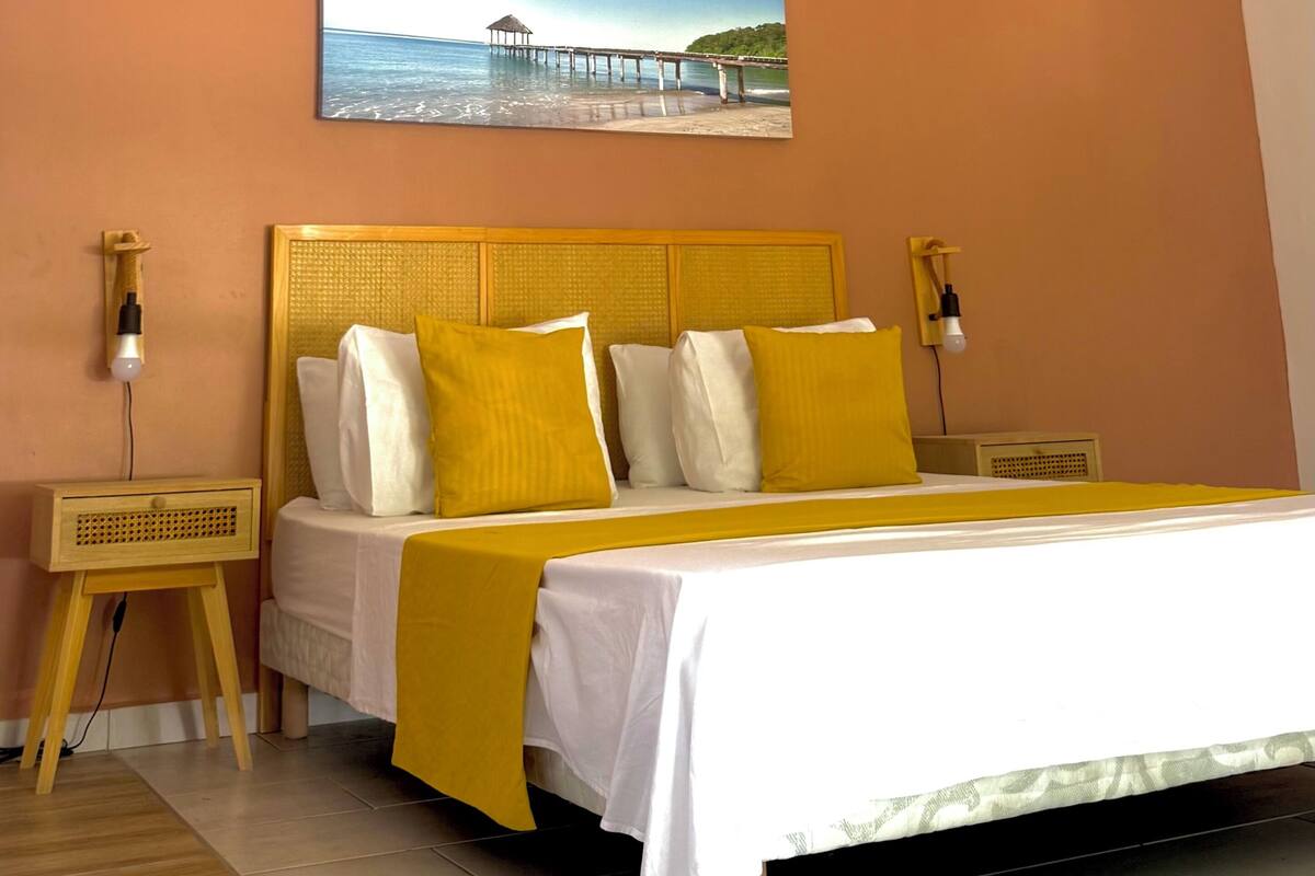 A bed with a white and yellow bedspread, two bedside tables with lamps, a painting of a pier on the wall, and a wooden floor.
