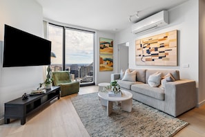Ein modernes Wohnzimmer mit einem grauen Sofa, einem schwarzen Fernseher, einem grünen Sessel, einem weißen Couchtisch und einem großen Fenster mit Blick auf die Stadt.