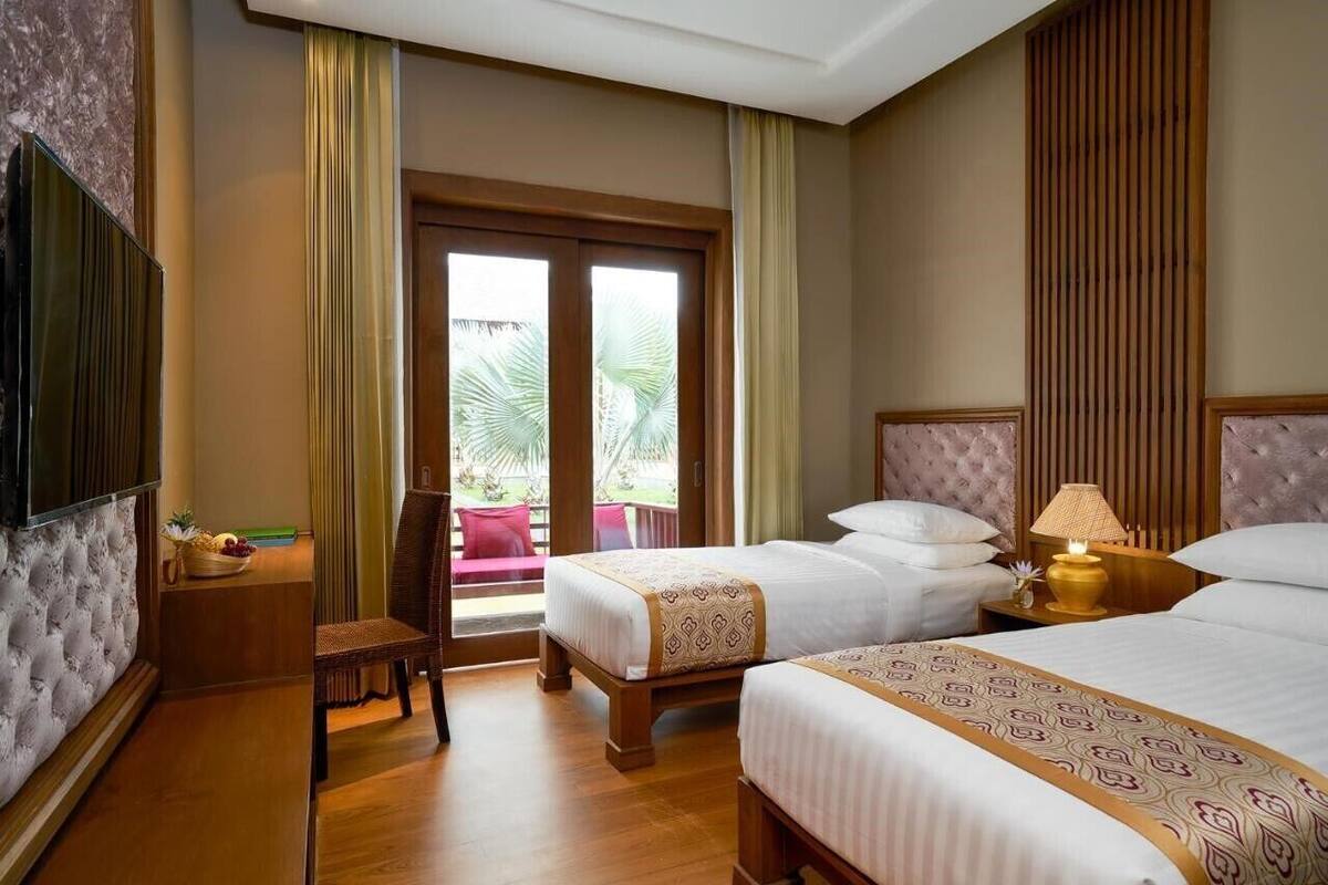 Habitación de hotel con dos camas, televisión, una mesita con frutero y vista a palmeras por la puerta.