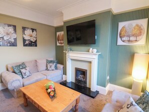 A living room with a sofa, a wooden coffee table with fruit, a fireplace, and wall-mounted artwork. - Crescent Bay House (Filey)