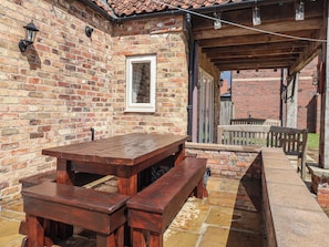 Eine Terrasse aus Ziegelsteinen mit einem Holztisch und Bänken, einem Fenster und einer Holzpergola.