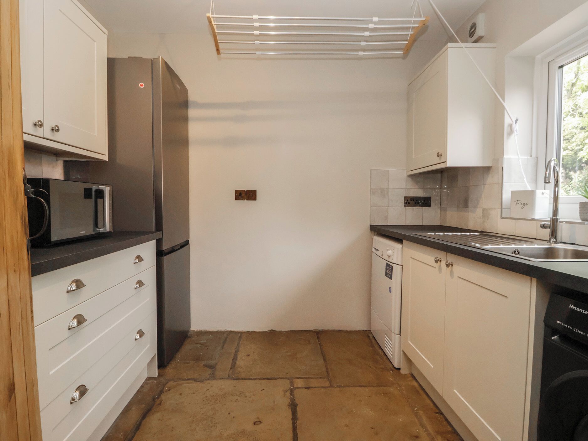 A compact kitchen with white cabinets, a stainless steel refrigerator, a microwave, a dishwasher, and a window with a view.