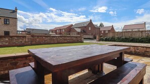 Une table de pique-nique en bois, avec des bancs, se trouve dans une cour bordée de bâtiments en briques.