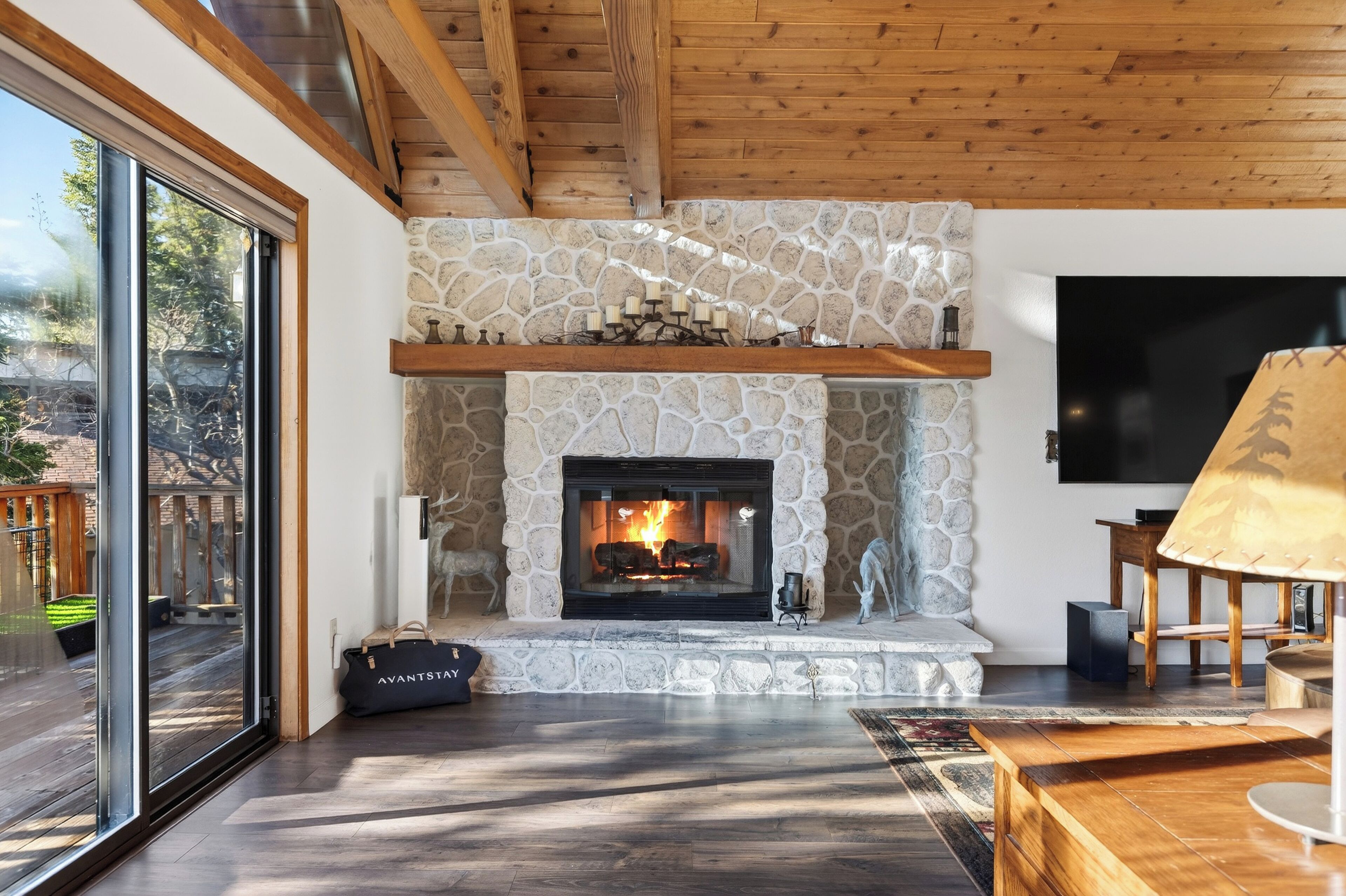 A modern living room with a stone fireplace, wooden beams, a large TV, and a sliding glass door to the outdoors.