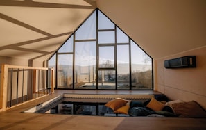 A high-ceilinged room with a large window, a wooden floor, and a sofa with cushions.