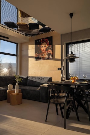 A modern living room with a sofa, dining table, and chairs, a large window, and a painting on the wall. - FlowFort View Private HotTub & Sauna Dom Swobodnie (Wojcieszyce)
