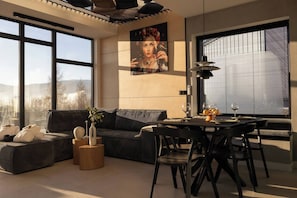 Ein modernes Wohnzimmer mit einer Couch, Esstisch und Stühlen, großen Fenstern und einem Gemälde an der Wand.