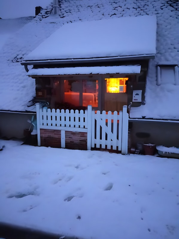 A snow-covered building with a white picket fence, a visible window, and a door leading to an interior with warm lighting.