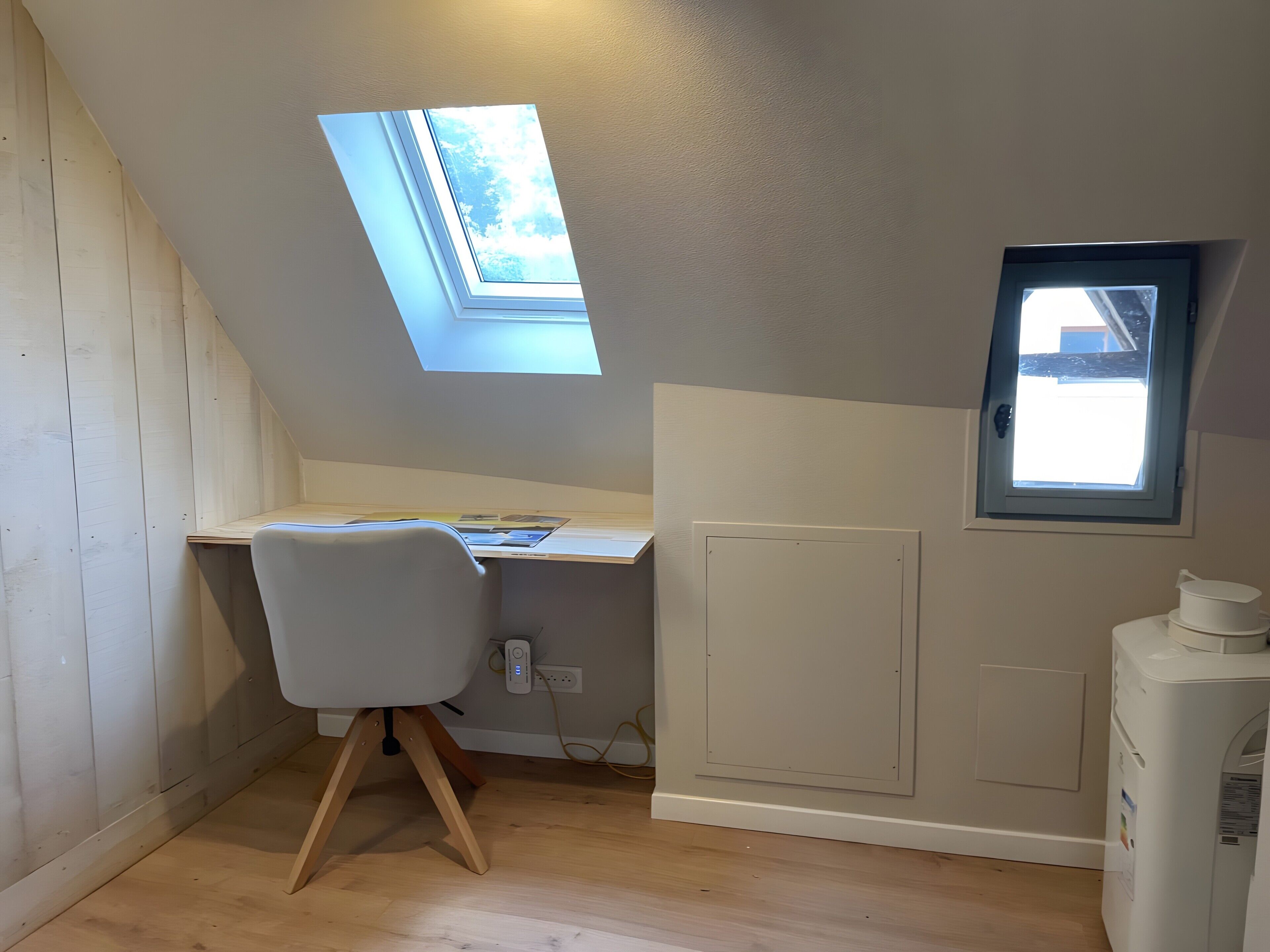 Une pièce avec un plafond en pente, un puits de lumière, un bureau, une chaise et un radiateur.