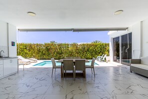 A modern kitchen with white cabinets, a dining area with a table and chairs, a pool, and a view of a lush garden. - Luxurious Villa with Private Pool in Sosua (Sosúa)