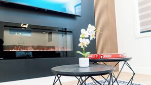 Ein modernes Wohnzimmer mit Fernseher, Kamin und einem kleinen Tisch mit einer Vasen voller weißer Orchideen.