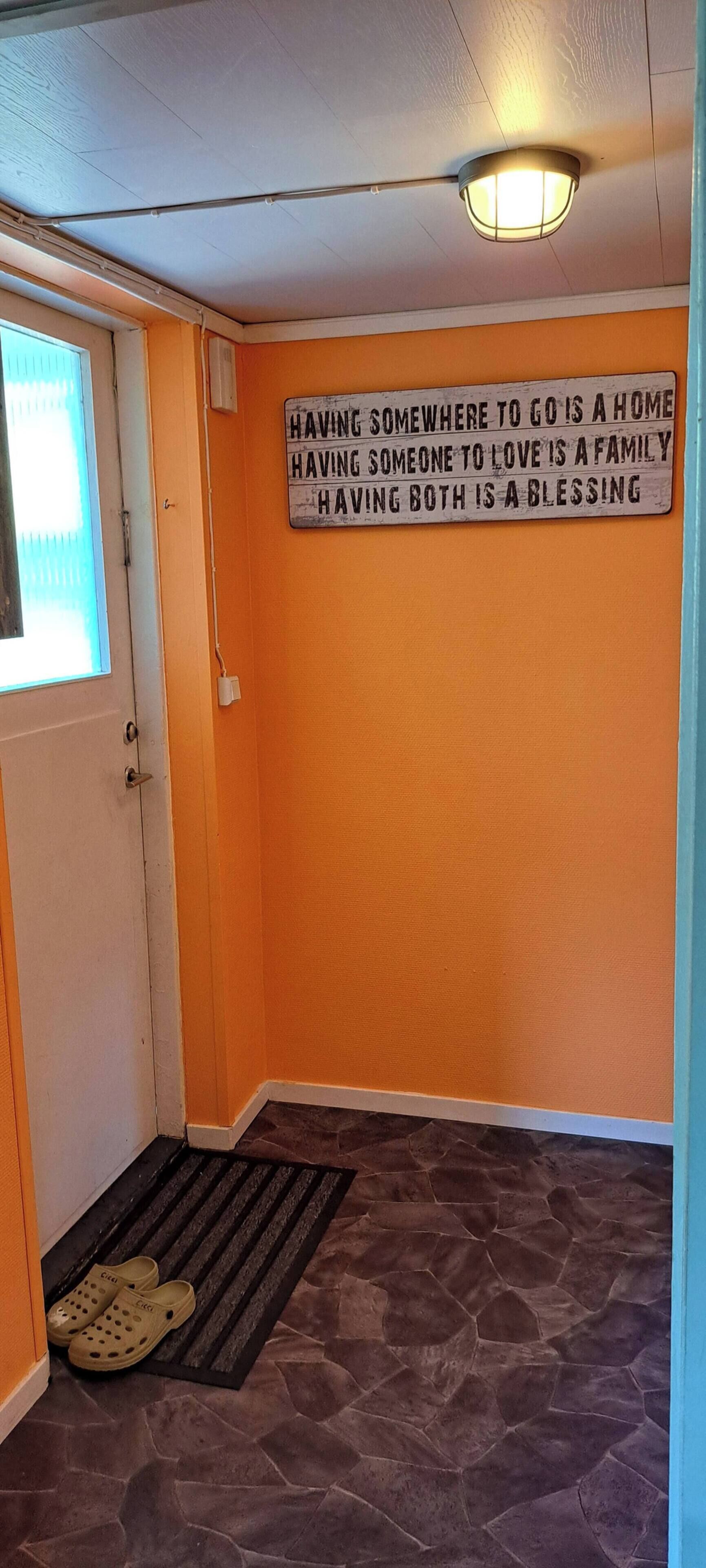 Un pasillo con pared naranja, puerta blanca y un cartel con un mensaje sobre el hogar y el amor familiar.