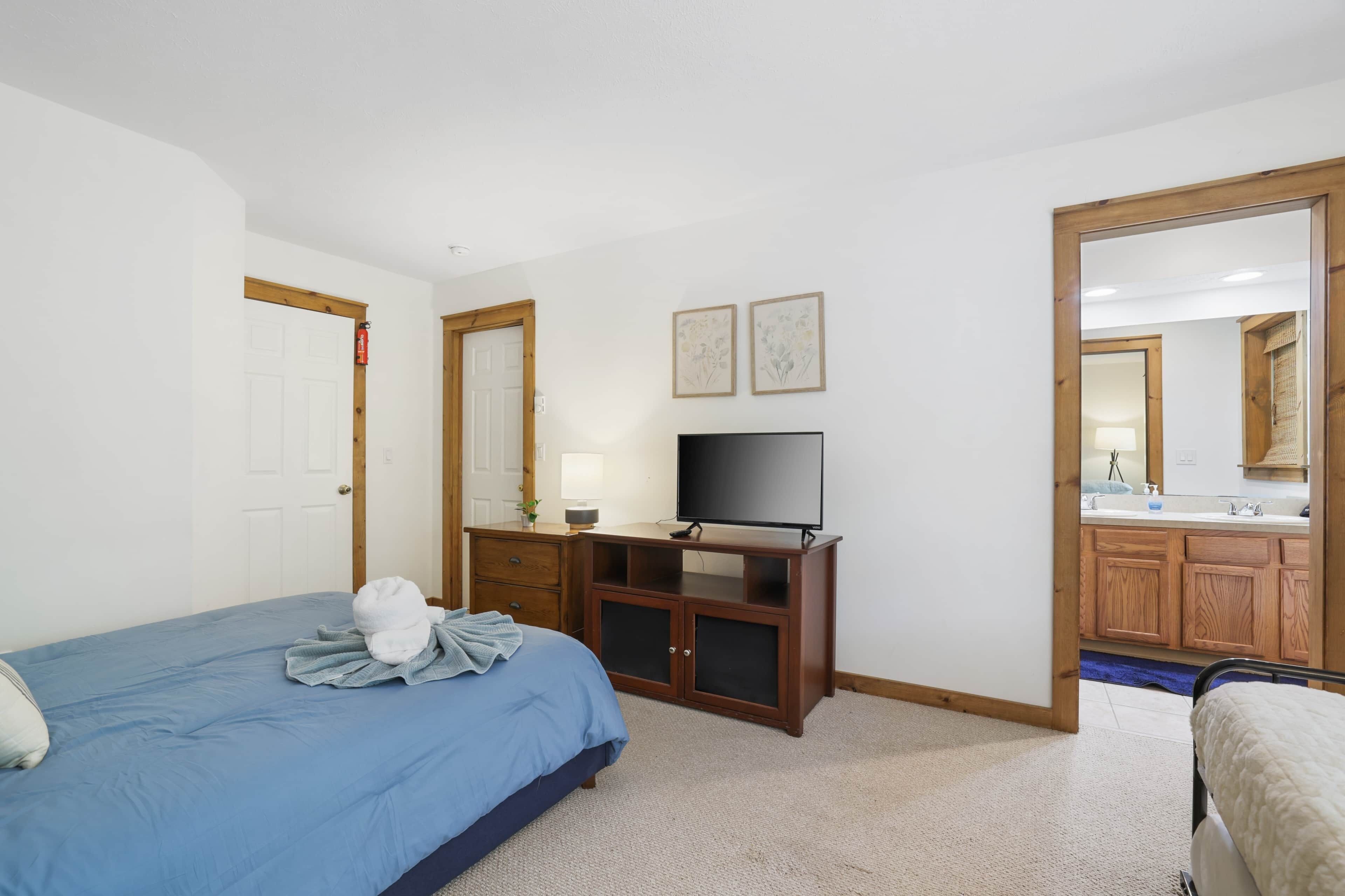 A hotel room with a bed, a TV, a desk, and a bathroom visible through an open door.