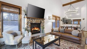 Una sala de estar con chimenea de piedra, un sofá, un sillón, una mesa de centro y una zona de cocina al fondo.