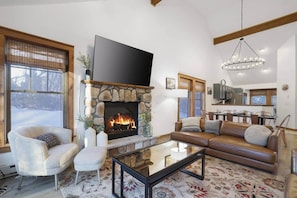 Una sala de estar con chimenea de piedra, un sofá, un sillón, una mesa de centro y una zona de cocina al fondo.
