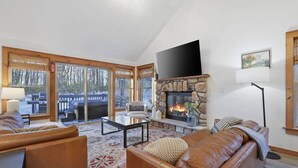 Una sala de estar acogedora con chimenea de piedra, sofás de cuero, un televisor de pantalla plana y ventanas amplias con vistas a una terraza.