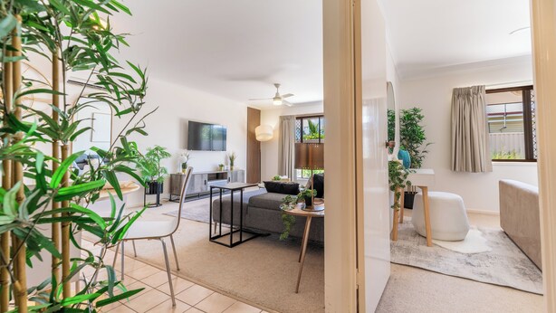 Een moderne woonkamer met een bank, salontafel en een eethoek met stoelen en een tafel. Er staan planten en er is een tv aan de muur bevestigd.