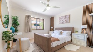 Een slaapkamer met een bed, nachtkastjes, een plafondventilator, een raam met gordijnen, een spiegel en een plant.