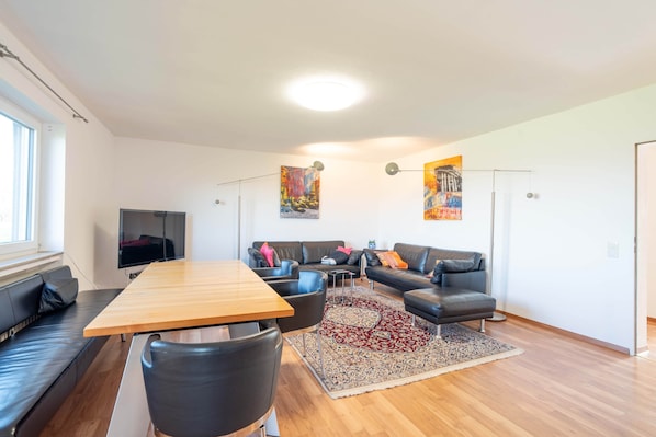 A modern living room with a wooden dining table, black leather sofas, a flat-screen TV, and a large rug.