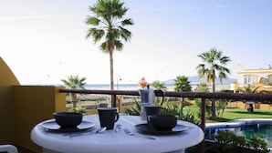 Ein Esstisch mit Schüsseln, Tassen und Besteck auf einem Balkon mit Blick auf einen Pool und Palmen.