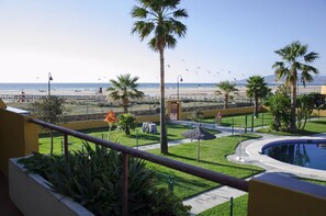 A balcony with a view of a beach, palm trees, a pool, and a fenced area with a lifeguard tower. - Apartamento Cosmico. (Tarifa)