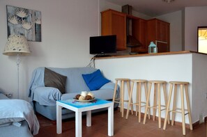 A living room with a sofa, a coffee table with decorative items, a TV, wooden stools, and a kitchen area with cabinets. - Apartamento Cosmico. (Tarifa)
