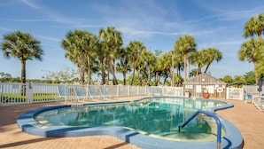 Ein Pool mit Backstein-Terrasse, umgeben von Palmen und einem eingezäunten Bereich mit Liegestühlen.