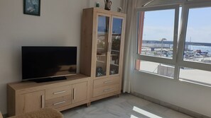 A room with a television on a wooden cabinet, a glass-fronted cabinet with decorative items, and a large window offering a view of a marina. - Puerto Bahía House . (Tarifa)