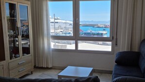 A room with a view of a marina, a white table, a sofa, and a cabinet with glass doors. - Puerto Bahía House . (Tarifa)