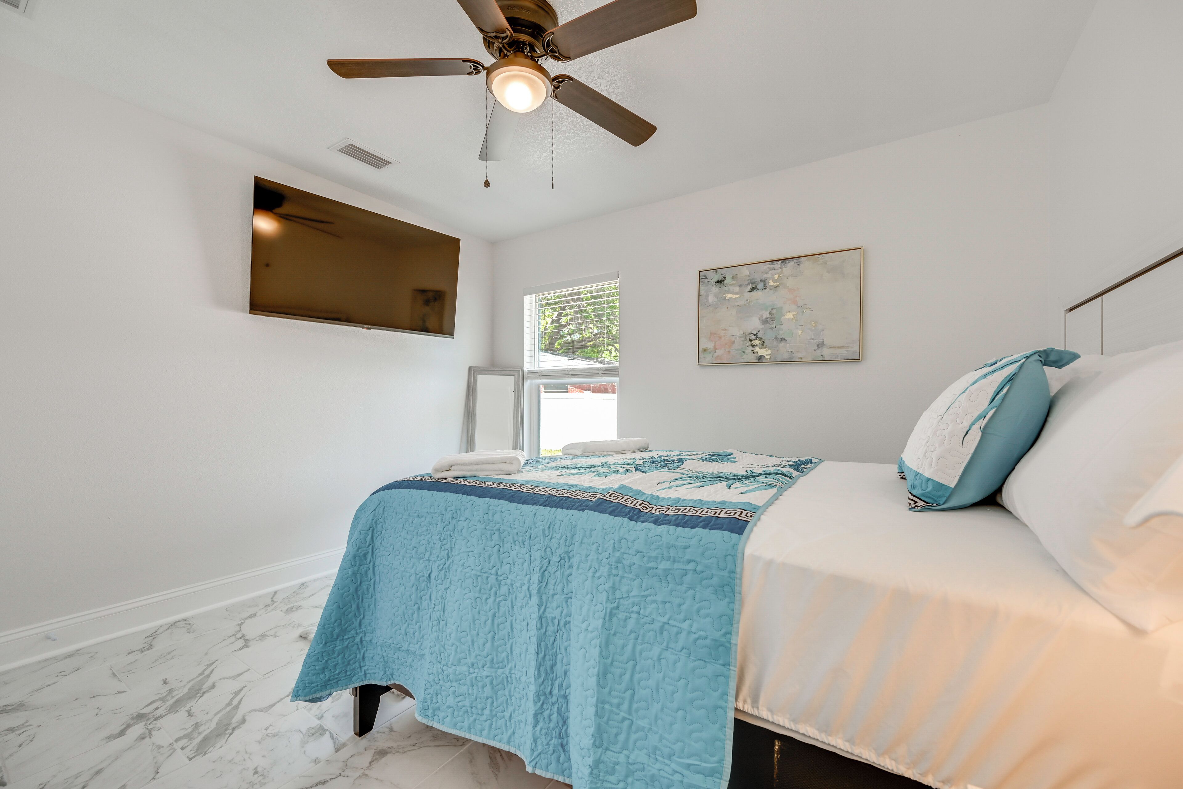 A bedroom with a bed, a ceiling fan, a window, and a painting on the wall.