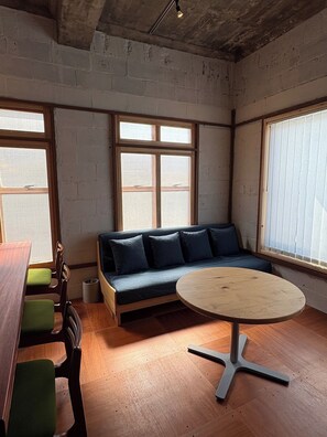 A room with a wooden table, a sofa with cushions, a window with blinds, and a concrete ceiling.