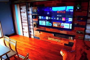 Una sala de estar moderna con una televisión de pantalla plana mostrando diferentes servicios de transmisión, una pared de acento de madera y una zona de comedor con mesa y sillas.