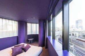 Een moderne hotelkamer met een bed, een klein tafeltje en uitzicht op de stad door grote ramen.