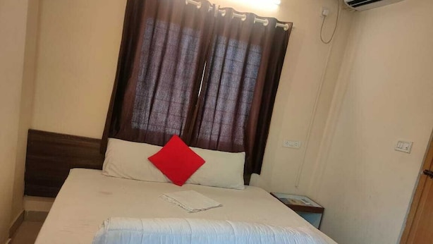 Una habitación de hotel con cama, mesita de noche, almohada roja y ventana con cortinas.