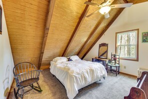 A bedroom with a sloped ceiling, a bed, a rocking chair, a small table, and a window with a view of trees.