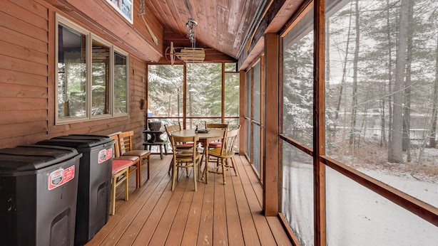 Eine Holzterrasse mit Tisch und Stühlen, einem Grill und einem großen Fenster mit Blick auf einen Wald.