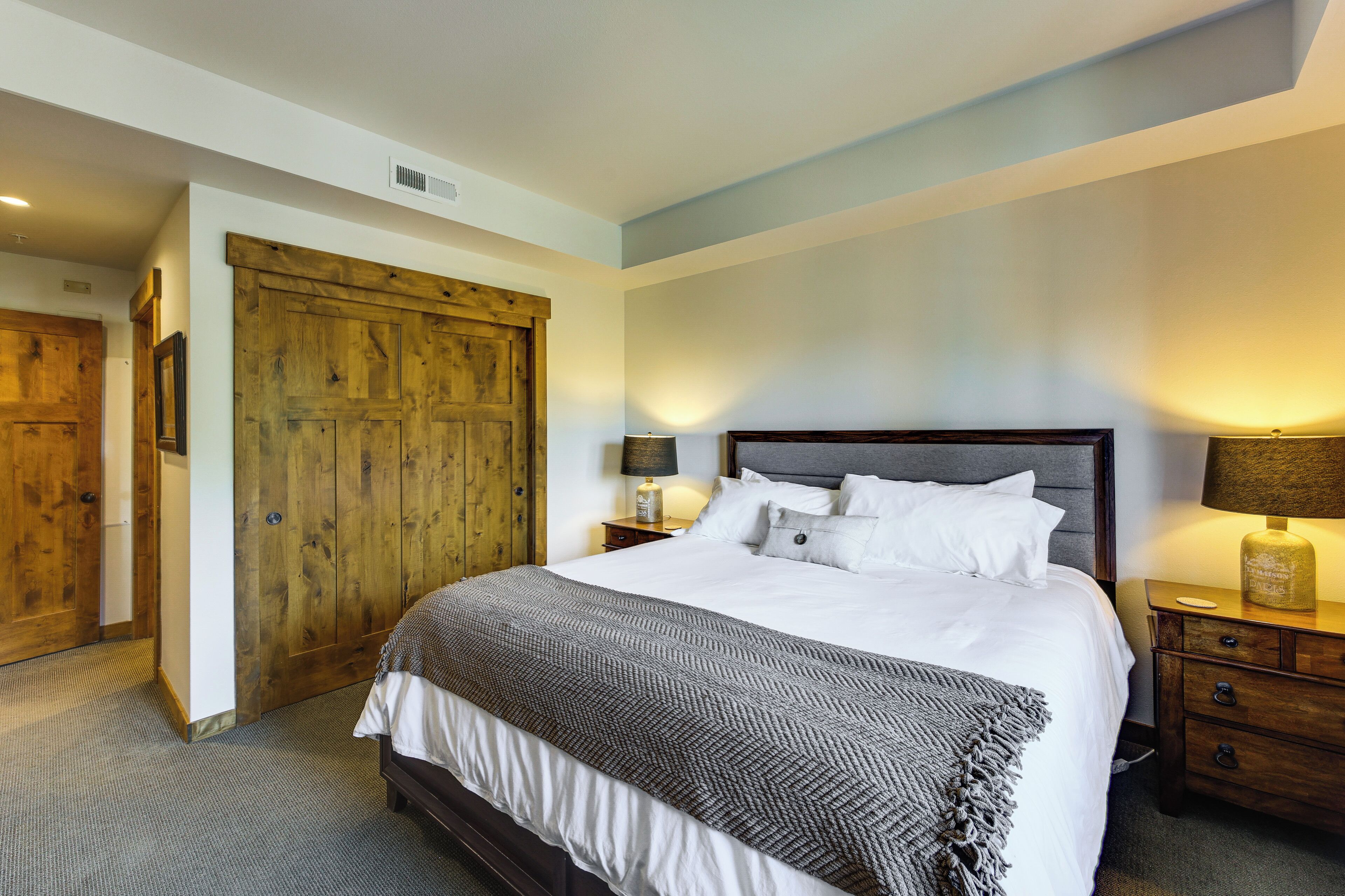Une chambre avec une grande armoire en bois, un lit recouvert de linge de maison blanc, deux tables de chevet avec des lampes et un sol recouvert d’une moquette.