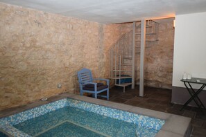 Una habitación con paredes de piedra, una piscina pequeña, una silla y una escalera en espiral de metal.