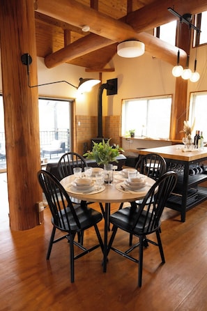 A dining area with a wooden table set for six, surrounded by black chairs, with a brick wall and windows in the background.