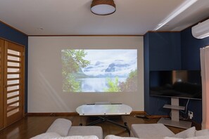 Ein Raum mit Projektionsleinwand, die eine Naturdarstellung zeigt, einem Flachbildfernseher, einer Holztür und einer Sitzecke mit Kissen.