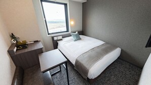 A compact hotel room with a bed, a small desk, a chair, a nightstand, a window with a view, and a wall-mounted lamp. - SUPER HOTEL Kirishima Natural Hot Spring (Kirishima)