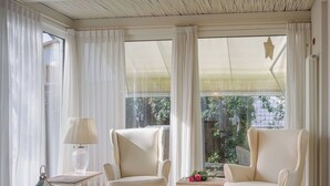 Ein Zimmer mit zwei Sesseln, einem kleinen Tisch, einer Lampe und Blick auf Grün hinter großen Fenstern.