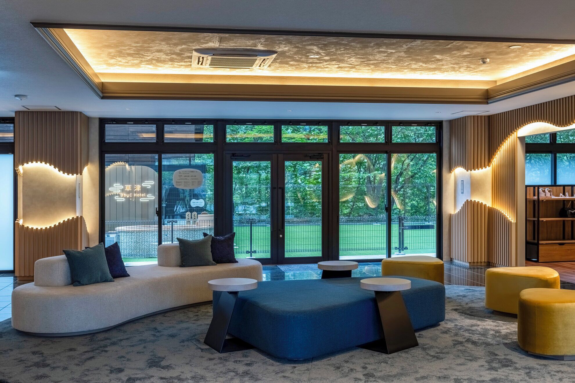 A modern hotel lobby with a curved sofa, round tables, and a large window offering a view of greenery.